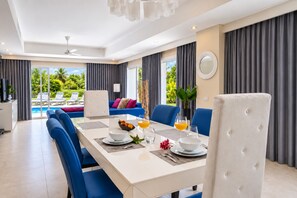A modern dining area with a white table set for two, blue chairs, and a view of a pool and greenery outside.