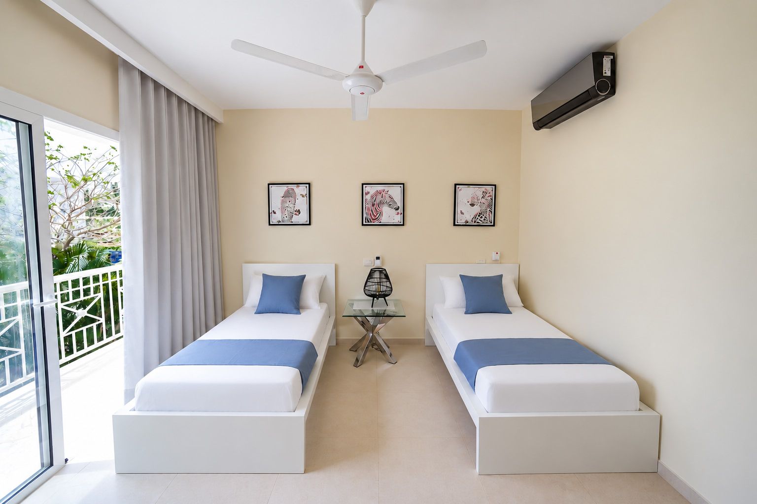Habitación de hotel con dos camas, un ventilador de techo, una mesita pequeña y cuadros en la pared.