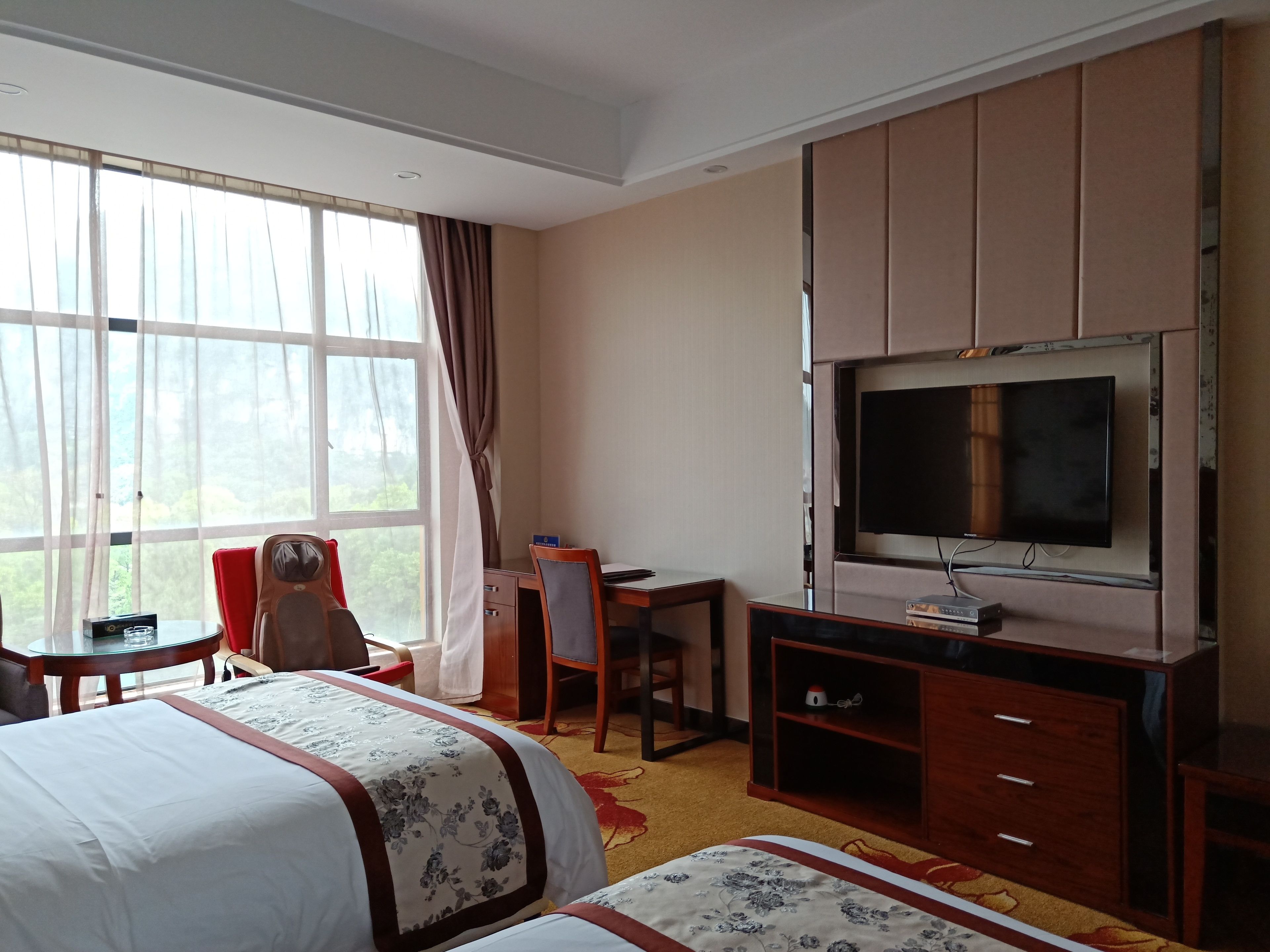 A hotel room with two beds, a desk, a chair, a television, and a large window with curtains.