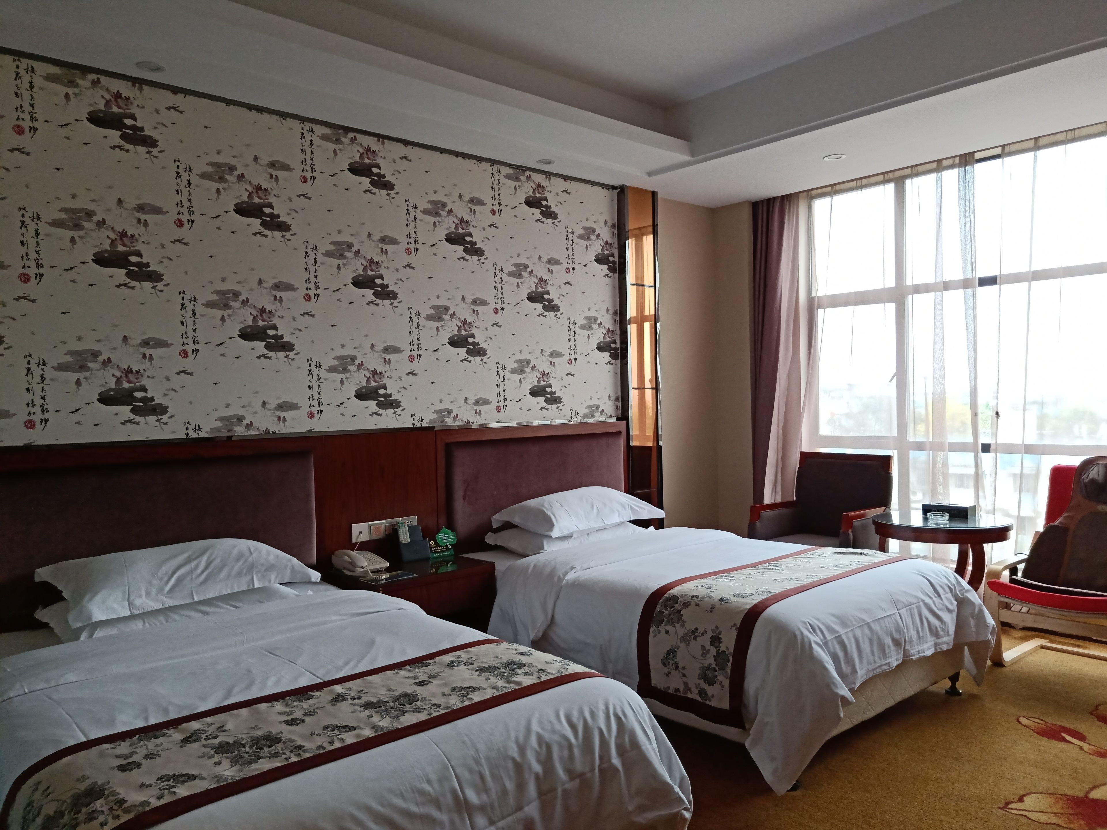 A hotel room with two beds, a wooden headboard, a large window with curtains, and a small table with a chair.