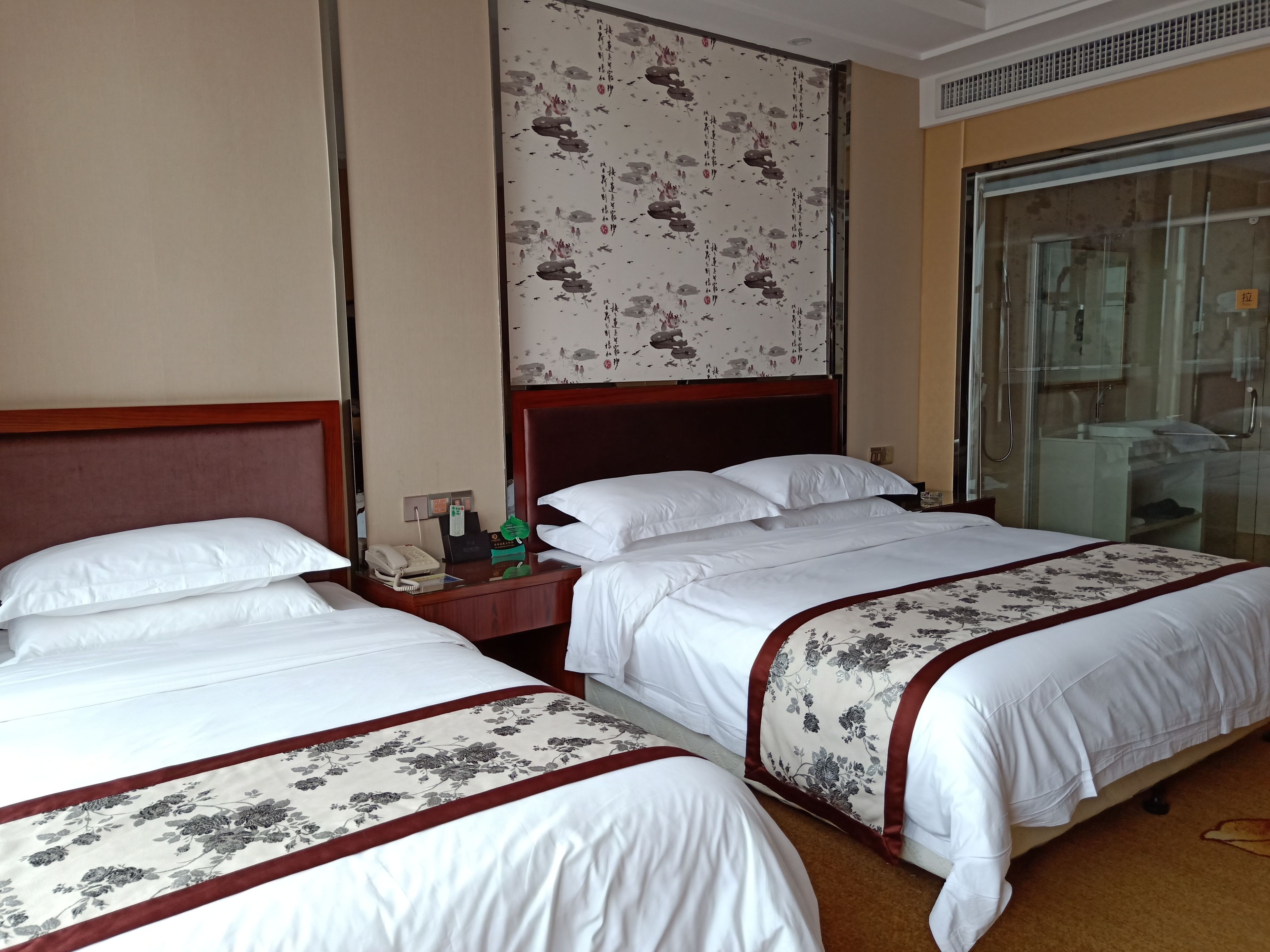 A hotel room with two beds, a wooden headboard, a nightstand with a phone, a decorative wall, and a glass shower area.
