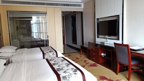 Une chambre d’hôtel avec deux lits, un bureau, une chaise, une télévision et une salle de bain visible à travers une porte en verre.