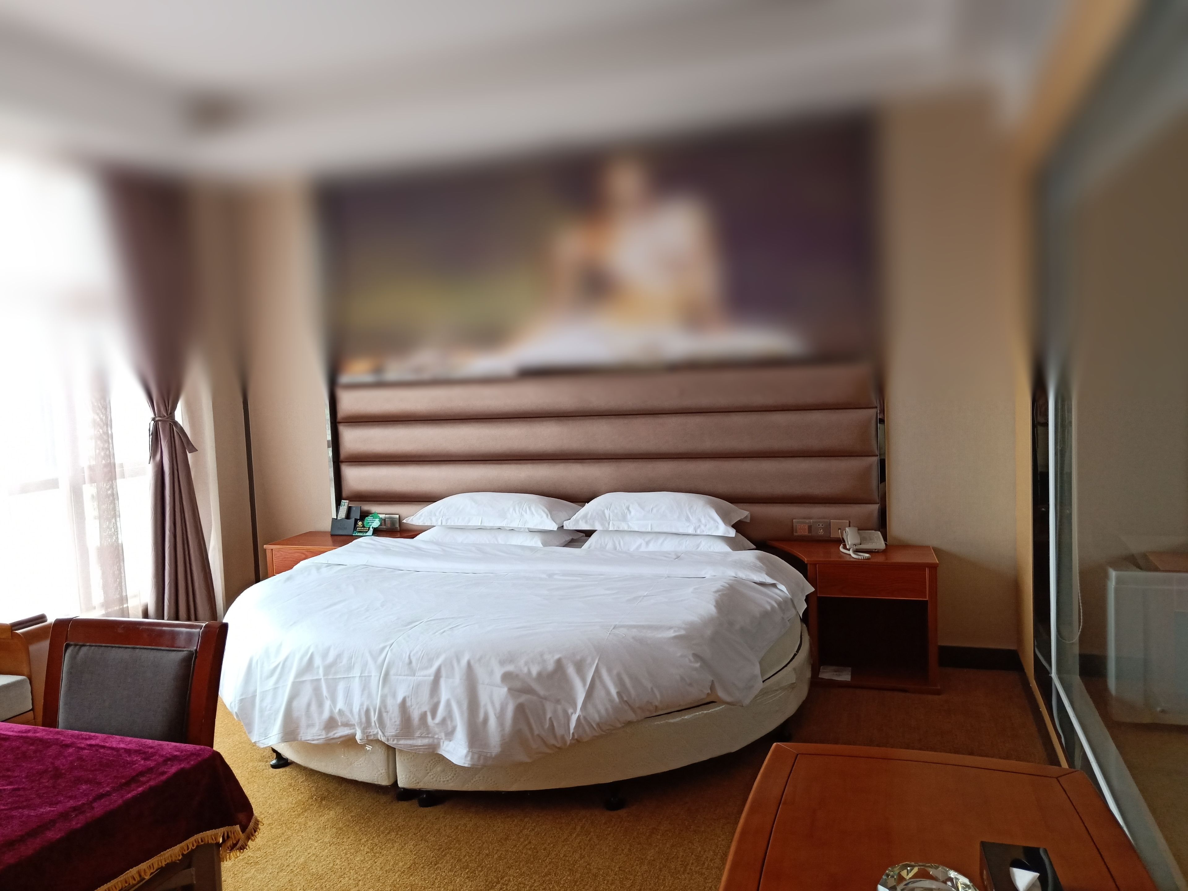 A hotel room with a round bed, a wooden nightstand, a desk, and a large window with curtains.