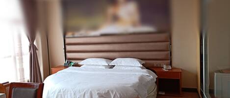 A hotel room with a round bed, a wooden nightstand, a desk, and a large window with curtains.