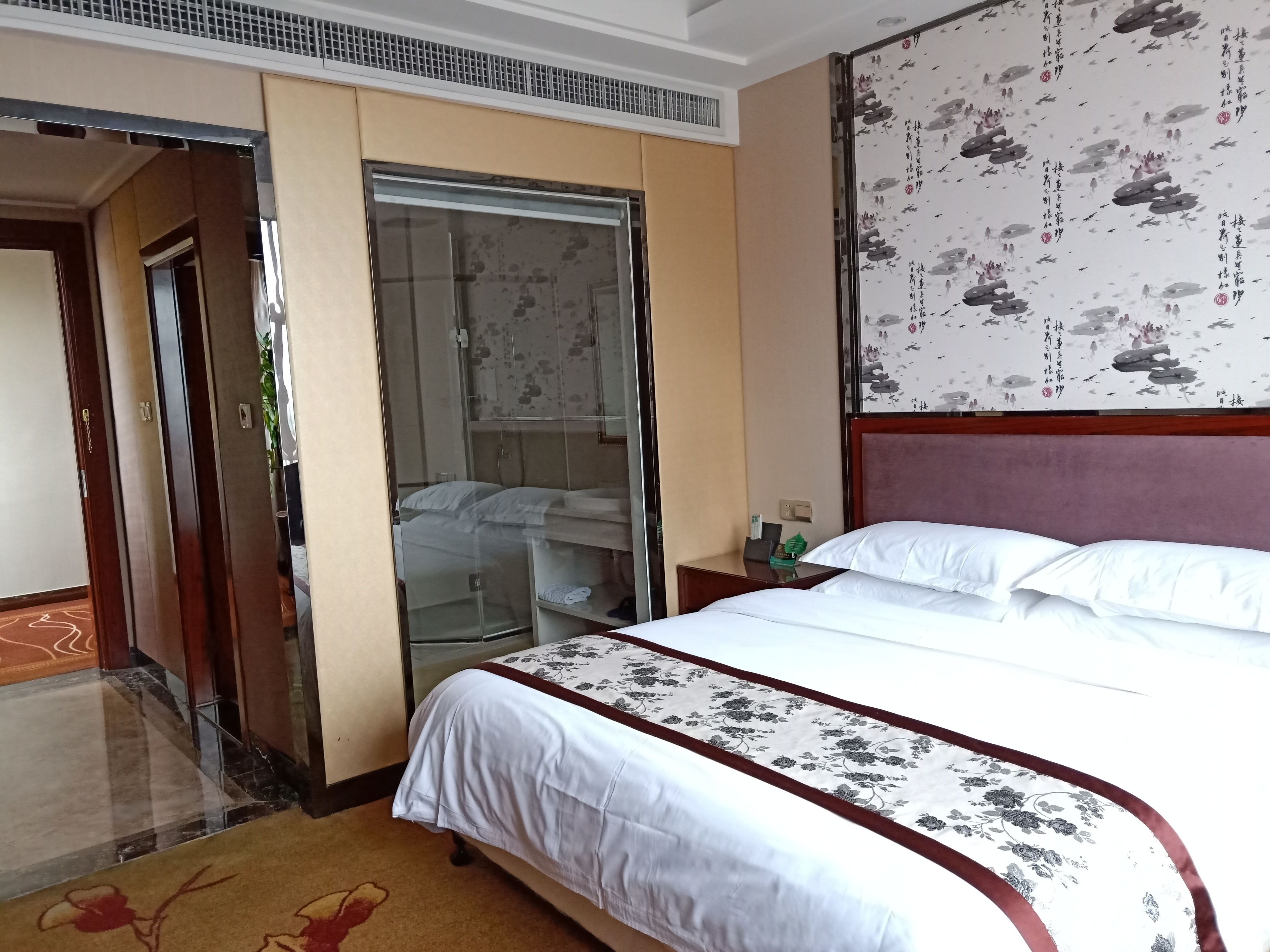 Une chambre d’hôtel avec un grand lit, une armoire, une salle de bain avec une porte en verre et un mur orné d’œuvres d’art traditionnelles.
