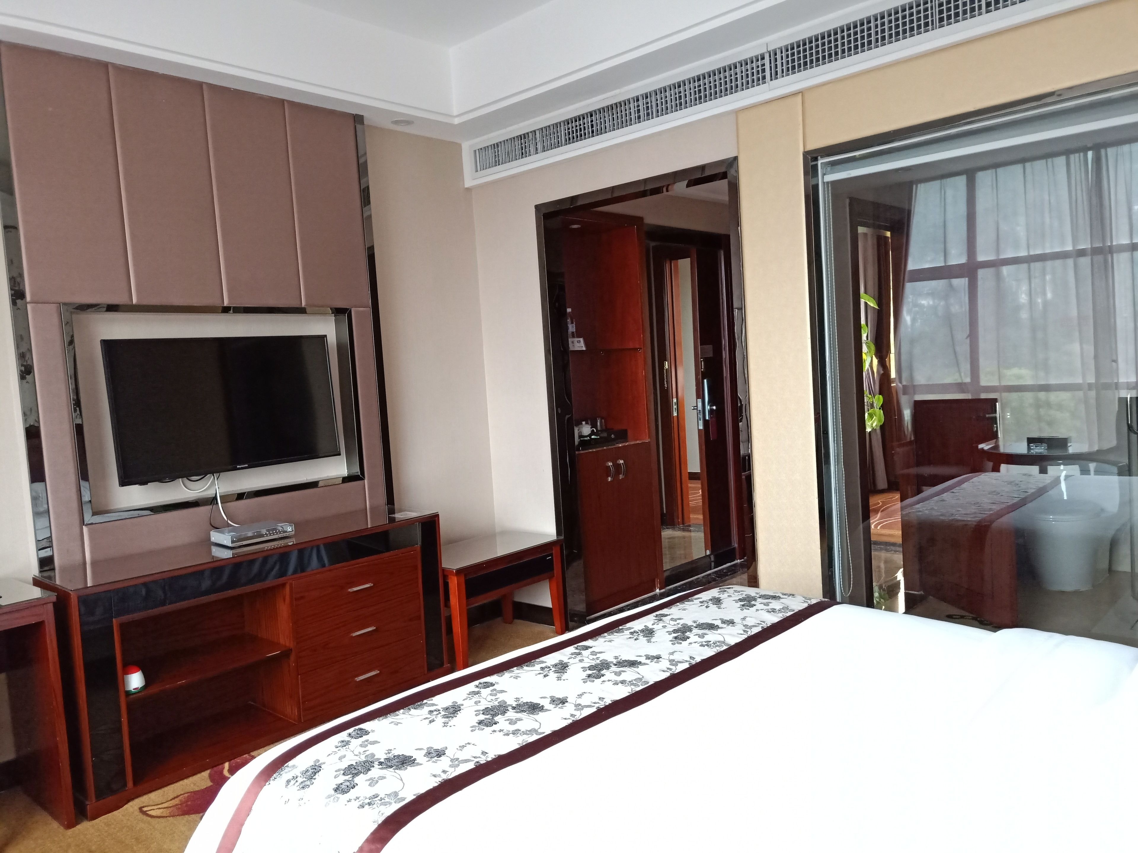 A hotel room with a bed, a TV on a stand, a wooden dresser, and a sliding glass door leading to a balcony.