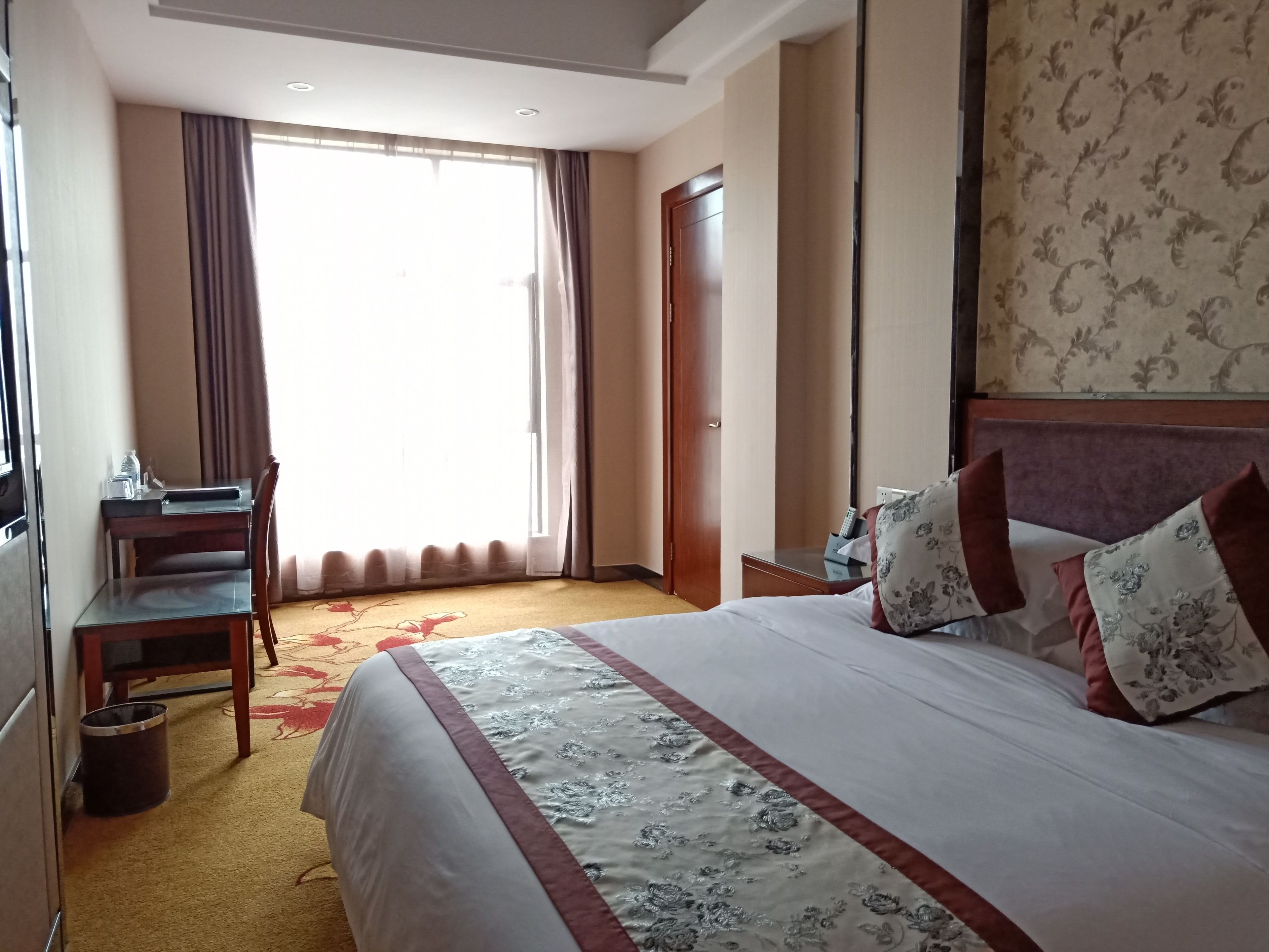 Habitación de hotel con una cama, un escritorio y una silla. Cuenta con una ventana con cortinas, una puerta y piso alfombrado.