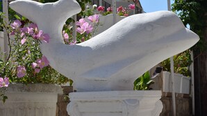 Eine weiße Steinstatue eines Vogels, der auf einer Säule sitzt, umgeben von Kübelpflanzen mit rosa Blüten.