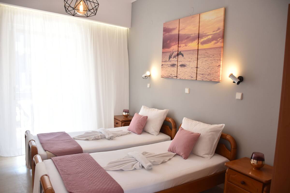 A hotel room with two beds, a large window with sheer curtains, a sunset painting on the wall, and a small table with a candle.