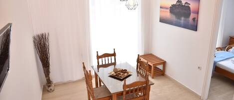 A dining area with a glass table, wooden chairs, a vase of dried branches, and a painting of a sunset over water.