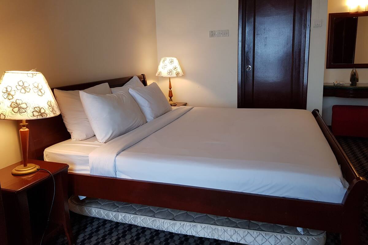 Een net opgemaakt bed met witte lakens en een houten hoofdbord, een bedlampje met een bloemendesign, en een donkerhouten deur.
