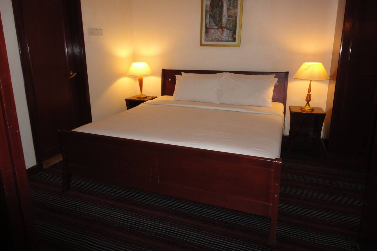 Een net opgemaakt bed met witte lakens, twee bedlampjes en een ingelijst kunstwerk boven het hoofdeinde.