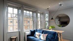 Un salon moderne avec un canapé bleu, une table en bois, un miroir rond et de grandes fenêtres donnant sur des immeubles.