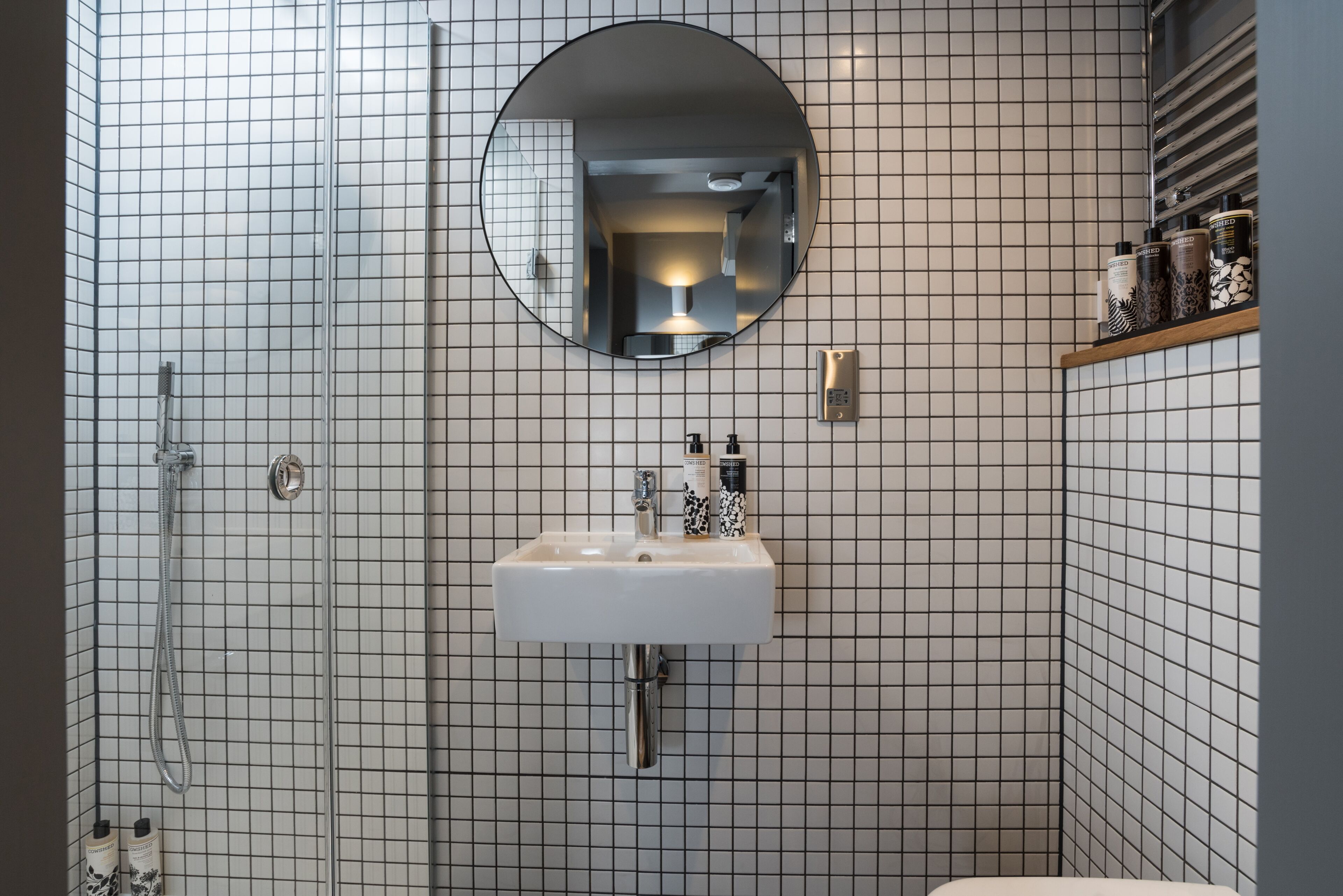 Une salle de bain avec un mur recouvert de carreaux blancs, un miroir rond, un lavabo avec un robinet et un espace douche.