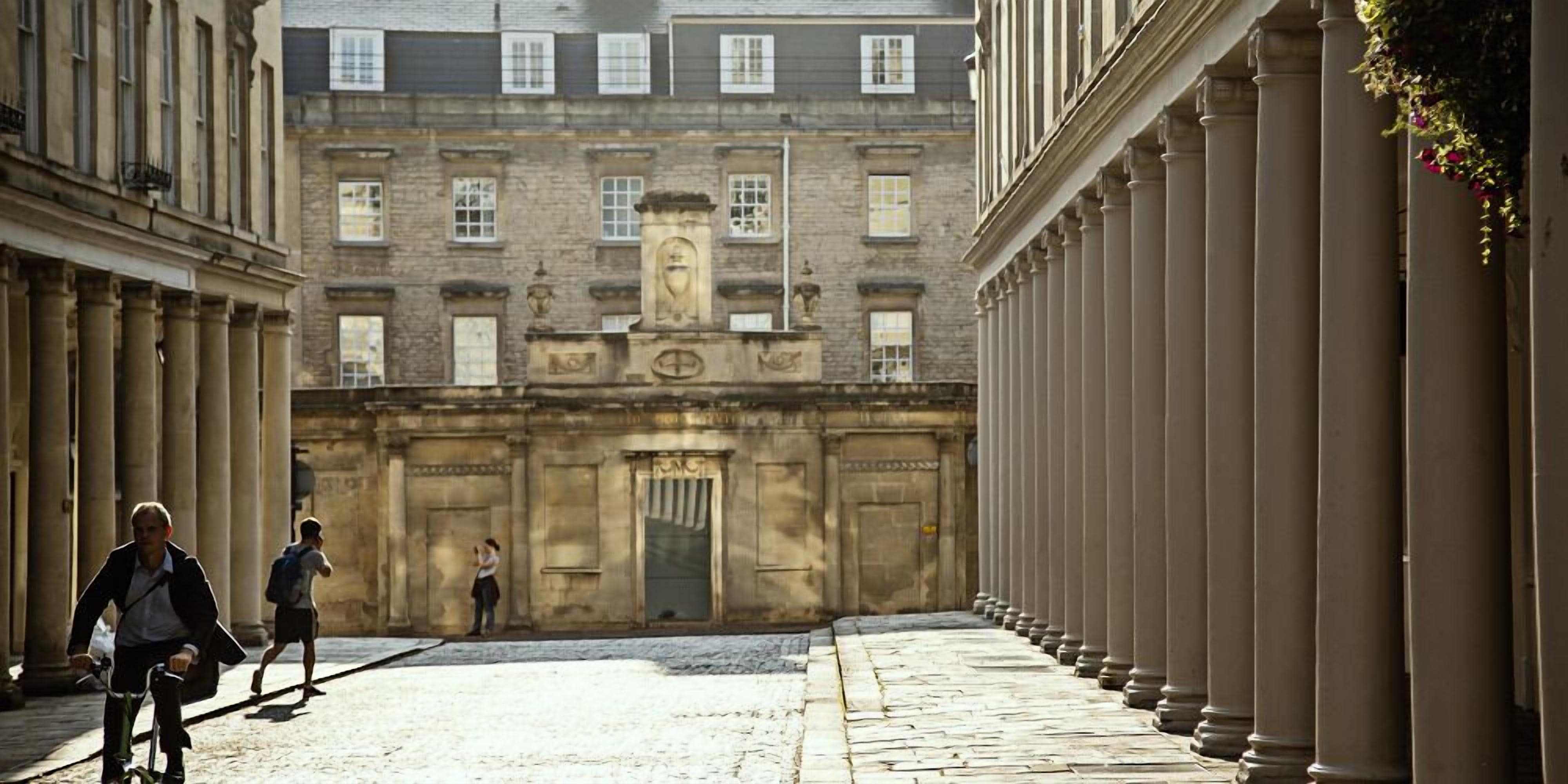 A narrow street with a building featuring classical architecture, including columns and a central entrance.