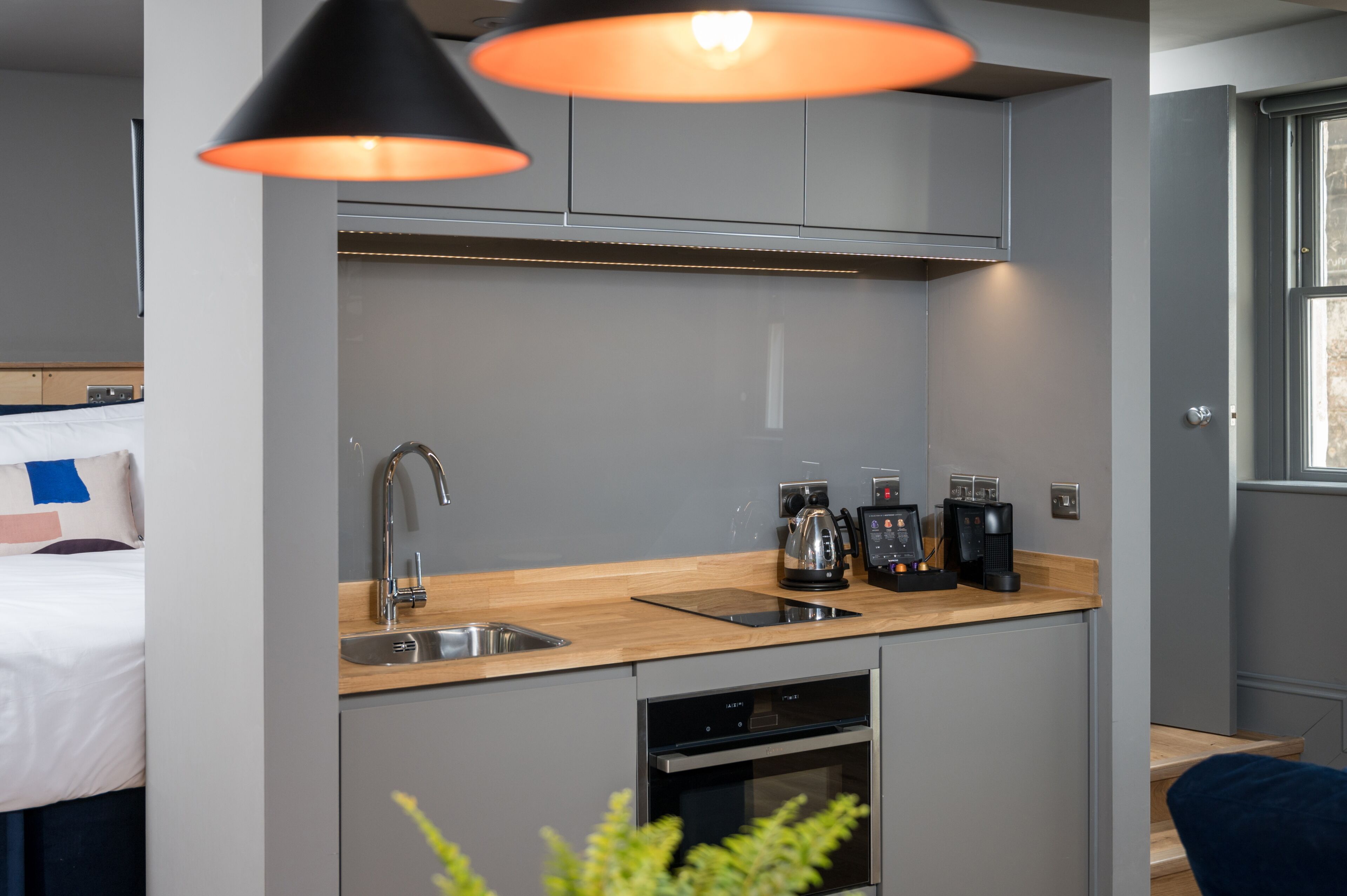 A compact kitchenette with a sink, stove, and coffee maker, a bed with a blue pillow, and a window with blinds.