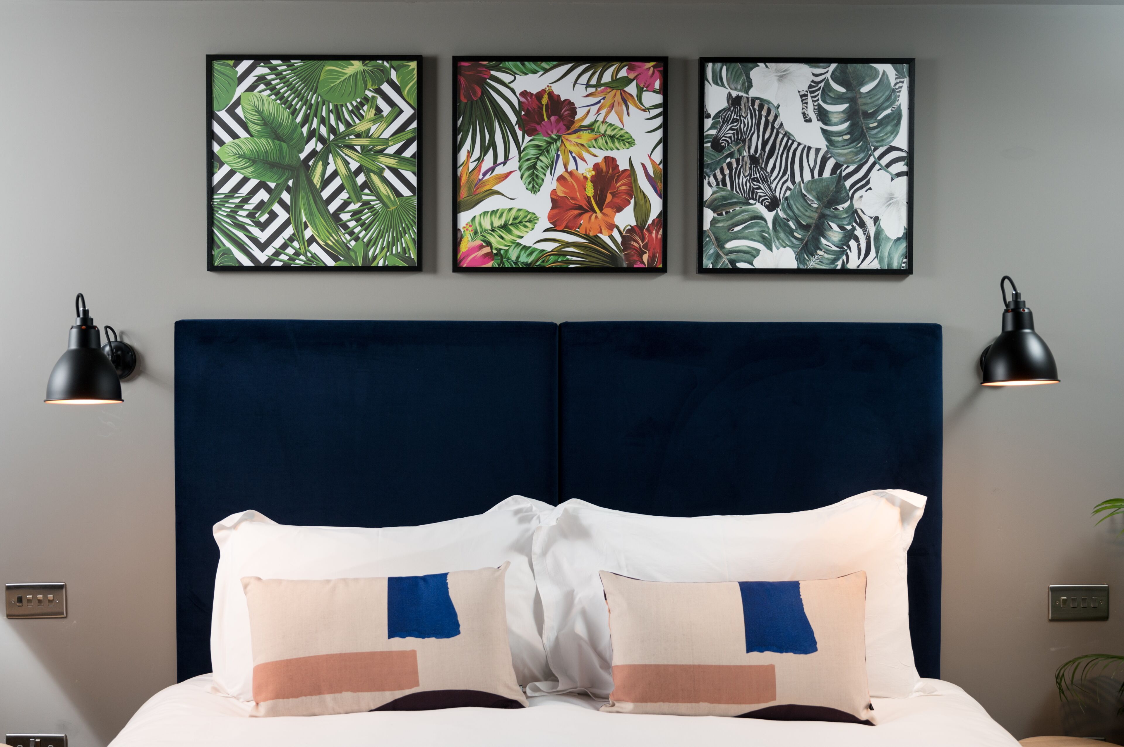 A bed with white and blue pillows, a dark blue headboard, and three framed tropical leaf paintings on the wall.