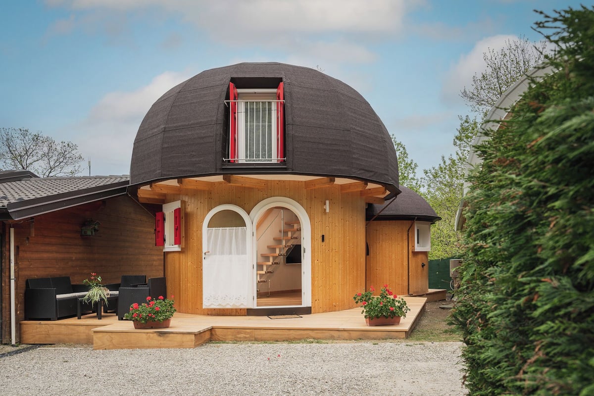 Una casa con forma de cúpula, fachada de madera, una puerta blanca y un pequeño porche con asientos y plantas en macetas.
