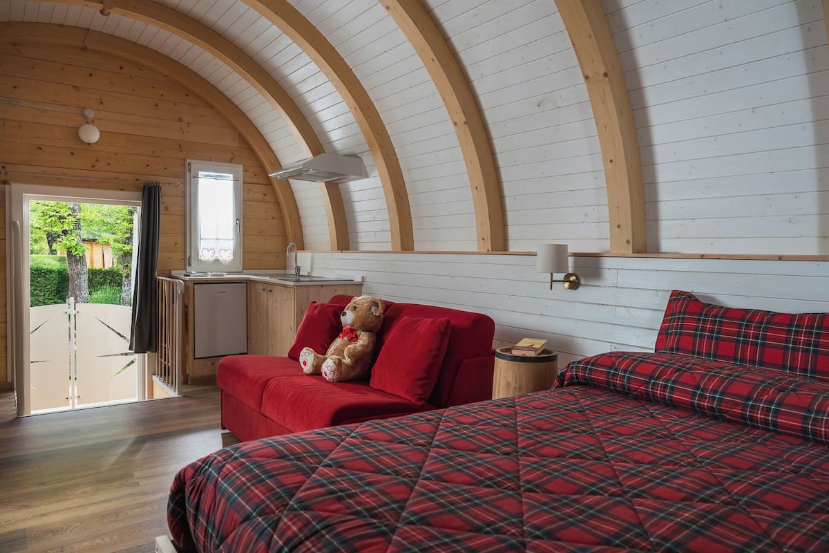 Una habitación acogedora con techo de madera, una cama con ropa de cama de cuadros rojos, un sofá rojo con un oso de peluche y una pequeña kitchenette.