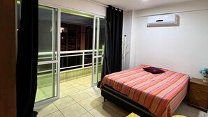 Un dormitorio con cama, un reposapiés amarillo, ventilador de techo y puerta corrediza de vidrio que da a un balcón.