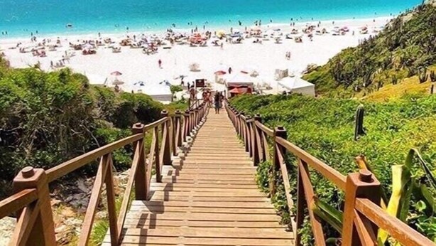 Una escalera de madera que lleva a una playa con mar azul cristalino y montañas al fondo.