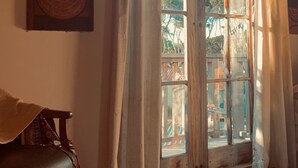 Ein Zimmer mit schrägem Dach, einem großen Fenster mit Vorhängen, einem Holzstuhl und einem an der Wand hängenden Kunstwerk.