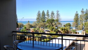 A balcony with a table and chairs, overlooking a coastal area with trees and parked cars. - Joanne Terrace 203 | Ocean Views & Kings Beach Escape (Caloundra)