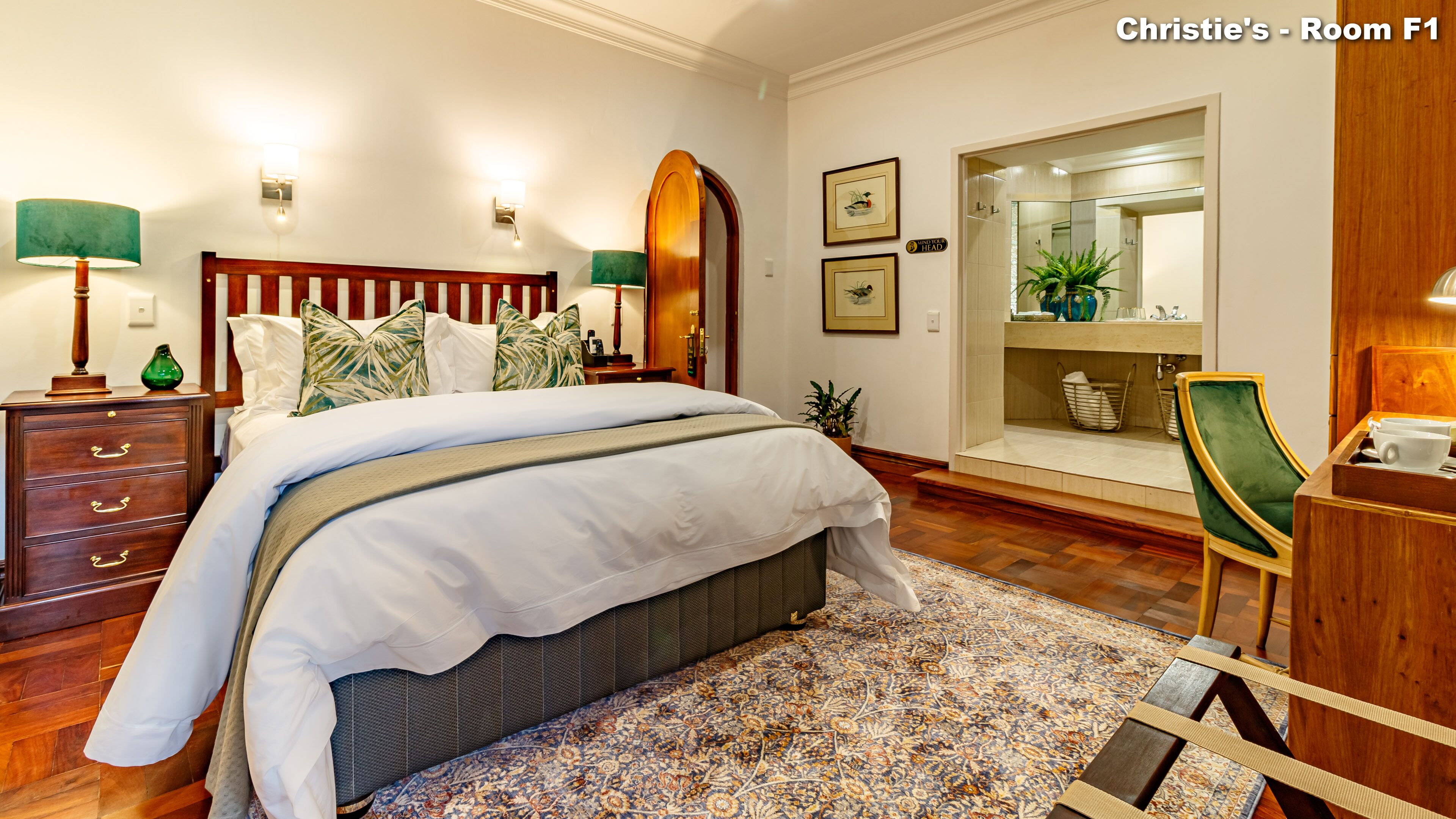 A neatly arranged bedroom with a large bed, a wooden dresser, a desk, and a bathroom visible through an arched doorway.