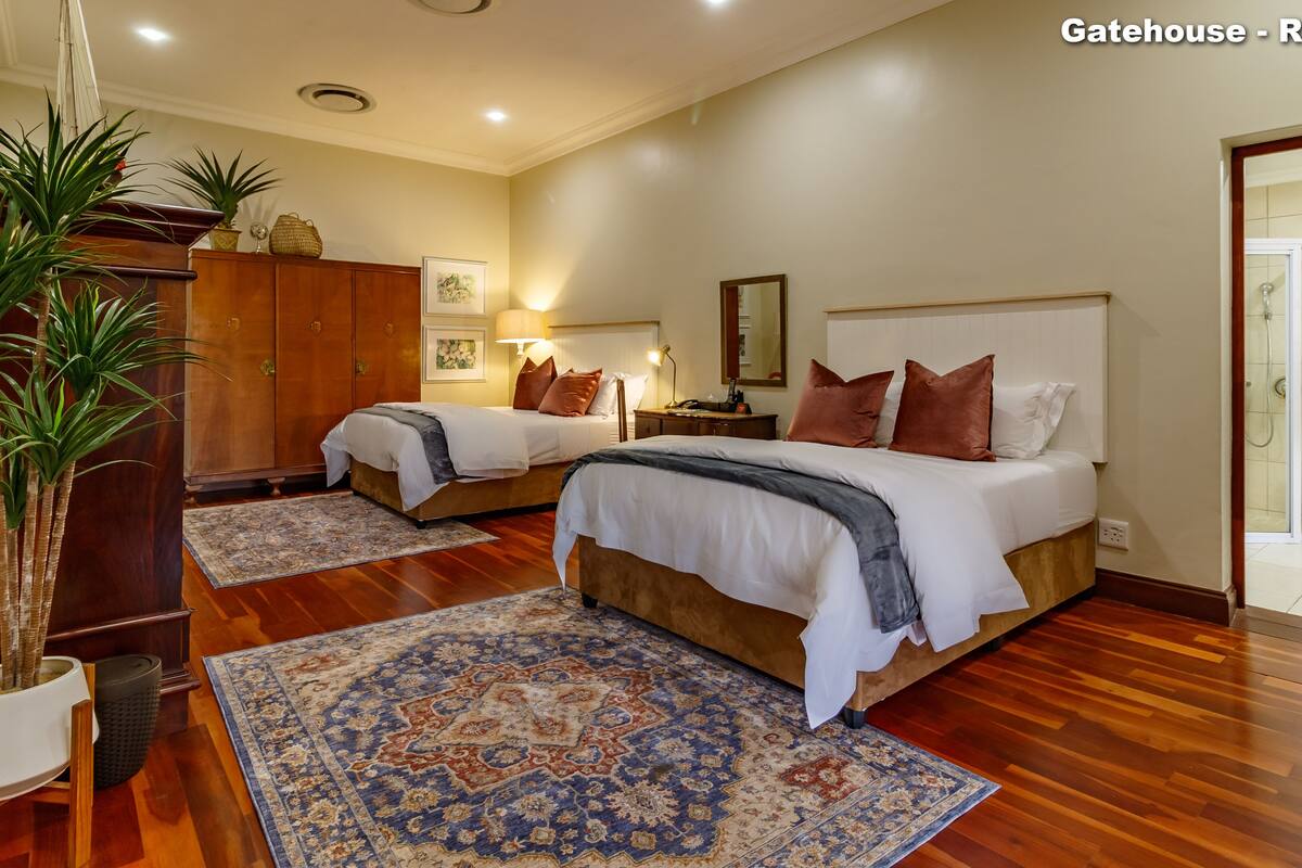 A hotel room with two beds, wooden flooring, a large rug, a potted plant, and a bathroom visible through an open door.