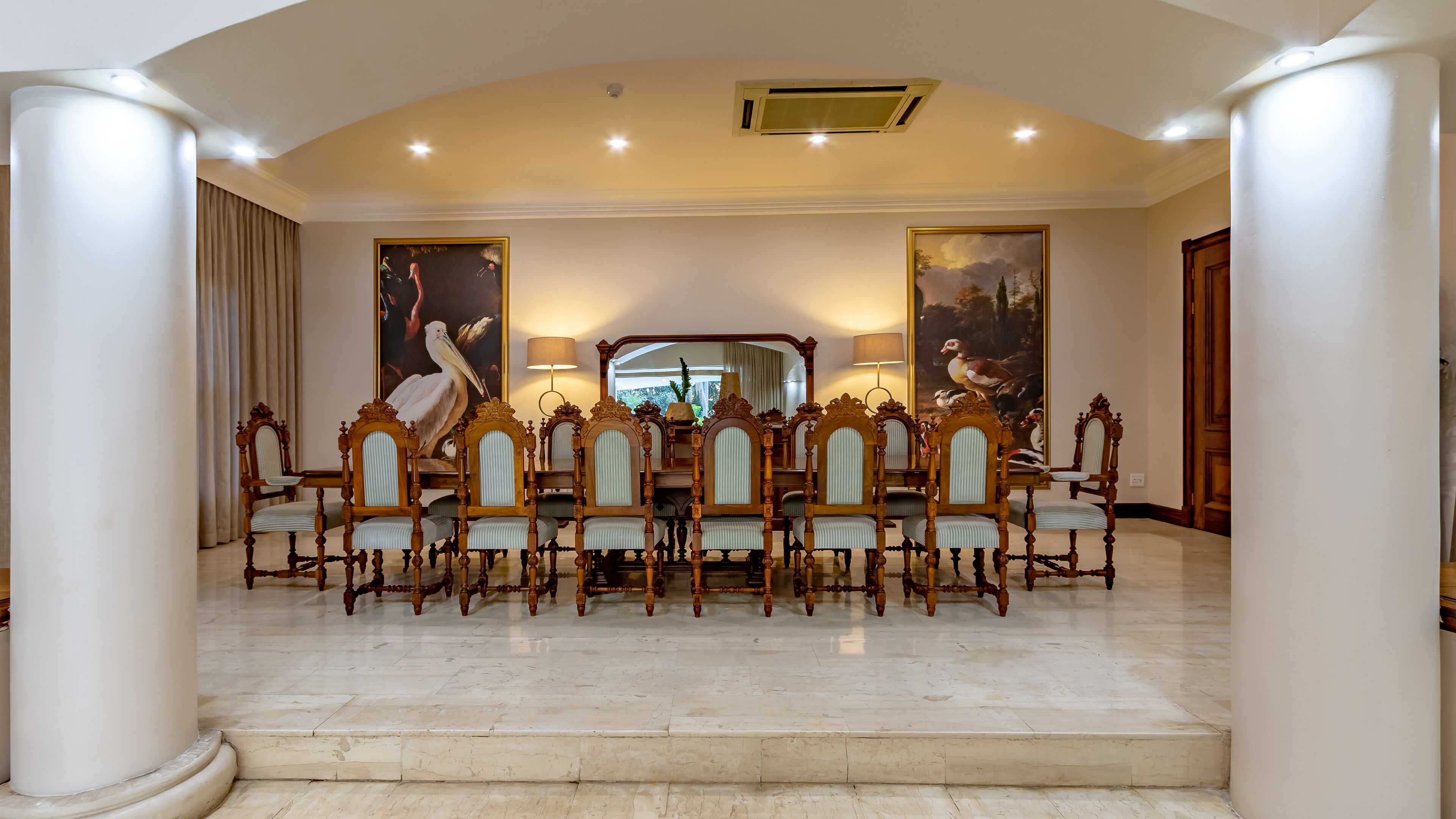 Un comedor formal con piso de mármol, sillas de madera ornamentadas y dos grandes cuadros en las paredes.