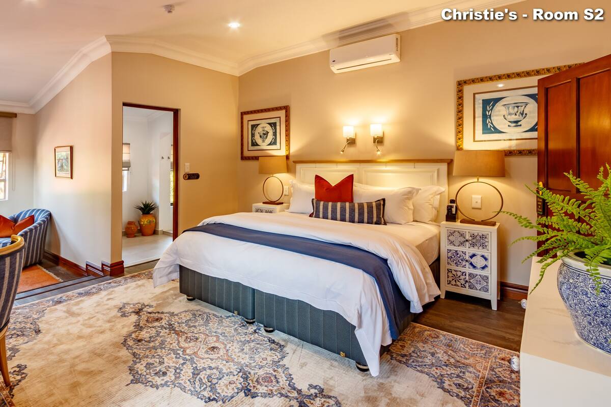 A well-lit bedroom with a large bed, bedside tables, a desk, and a chair. There is a window with a view, a door, and a wall-mounted air conditioner.