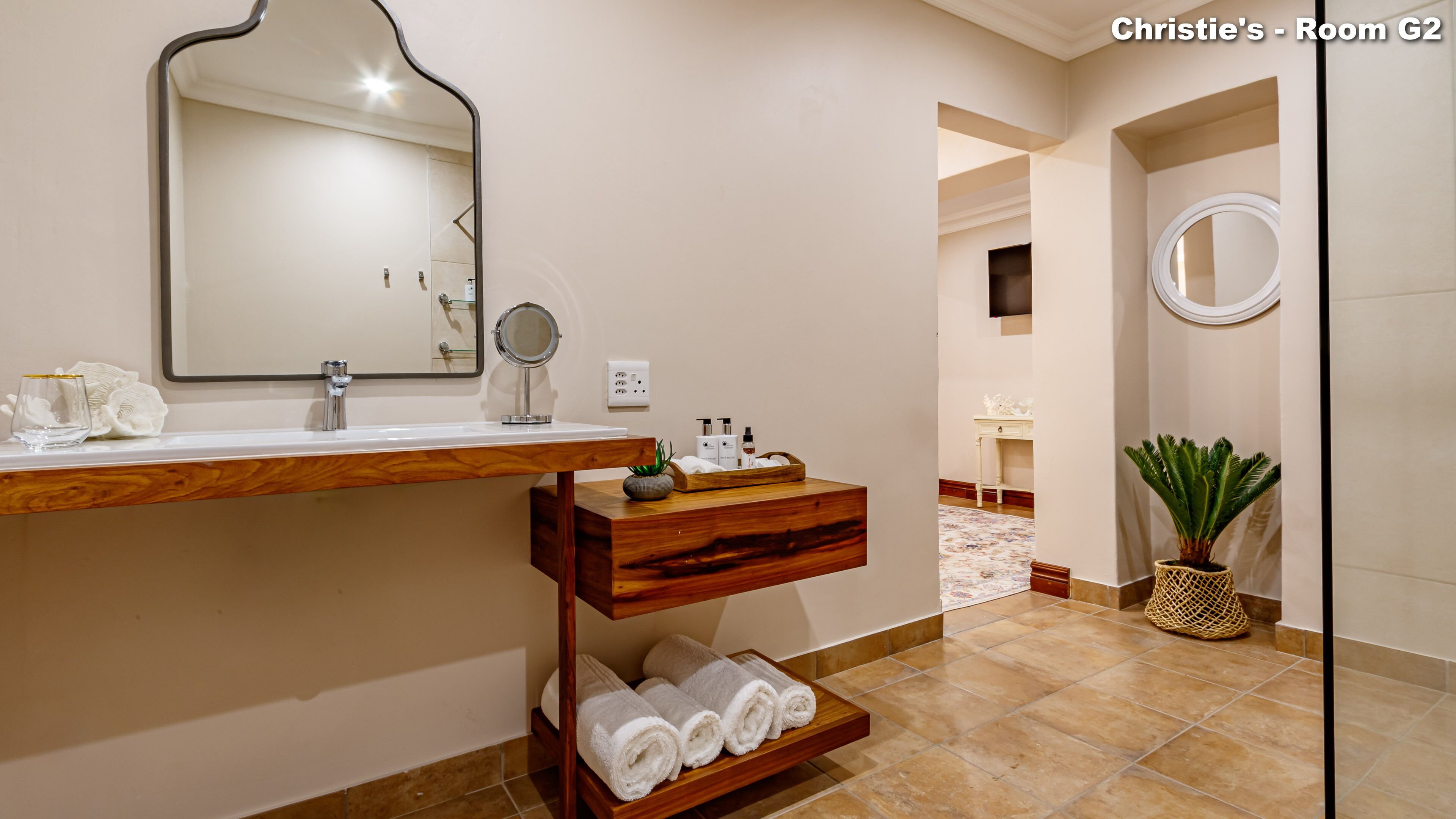 A bathroom with a wooden vanity, a large mirror, a small round mirror, and a shelf with toiletries. There are towels on a wooden shelf and a potted plant.