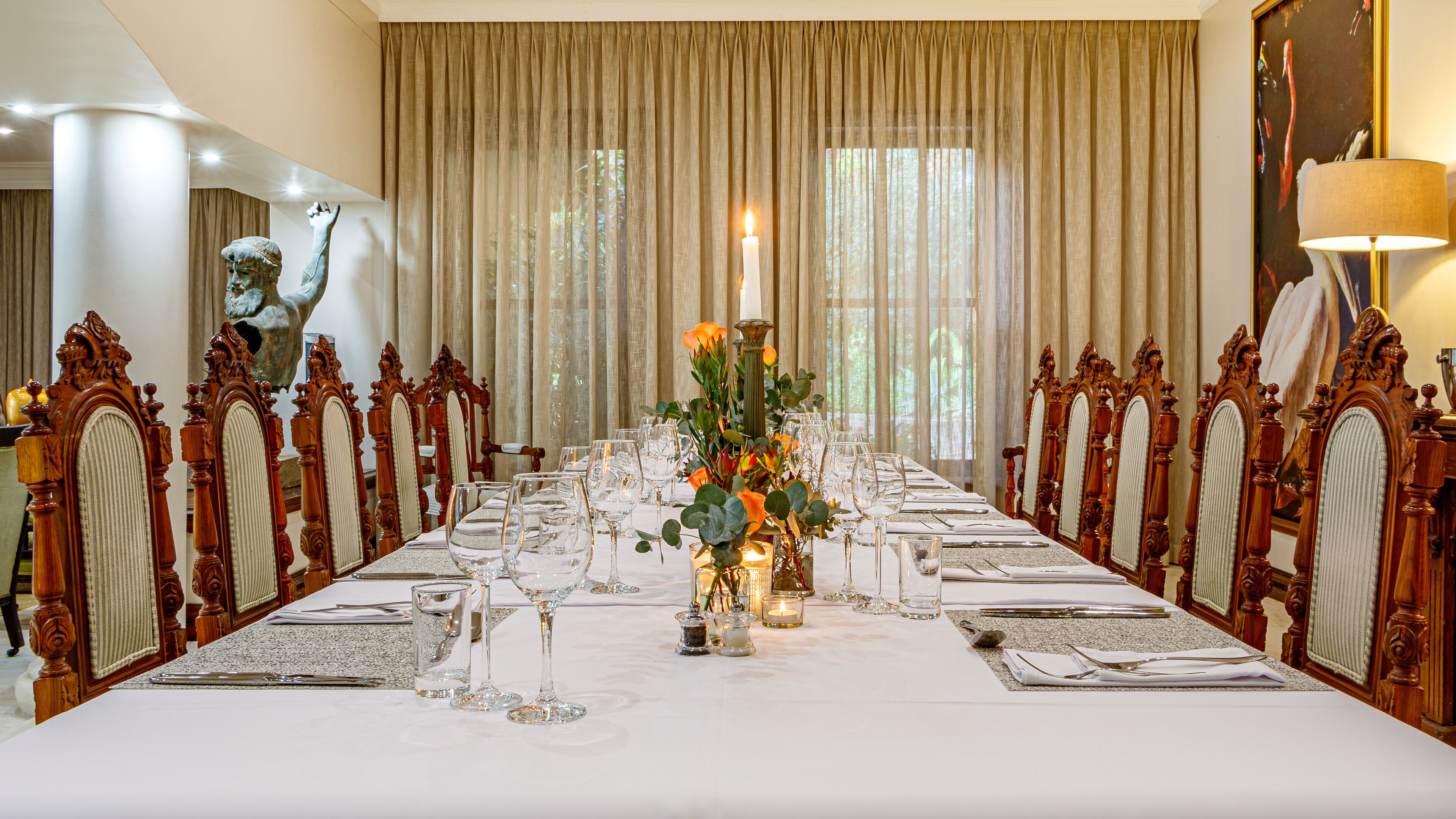 A formal dining room with a long table set for a meal, flanked by ornate chairs, and a statue on the left.