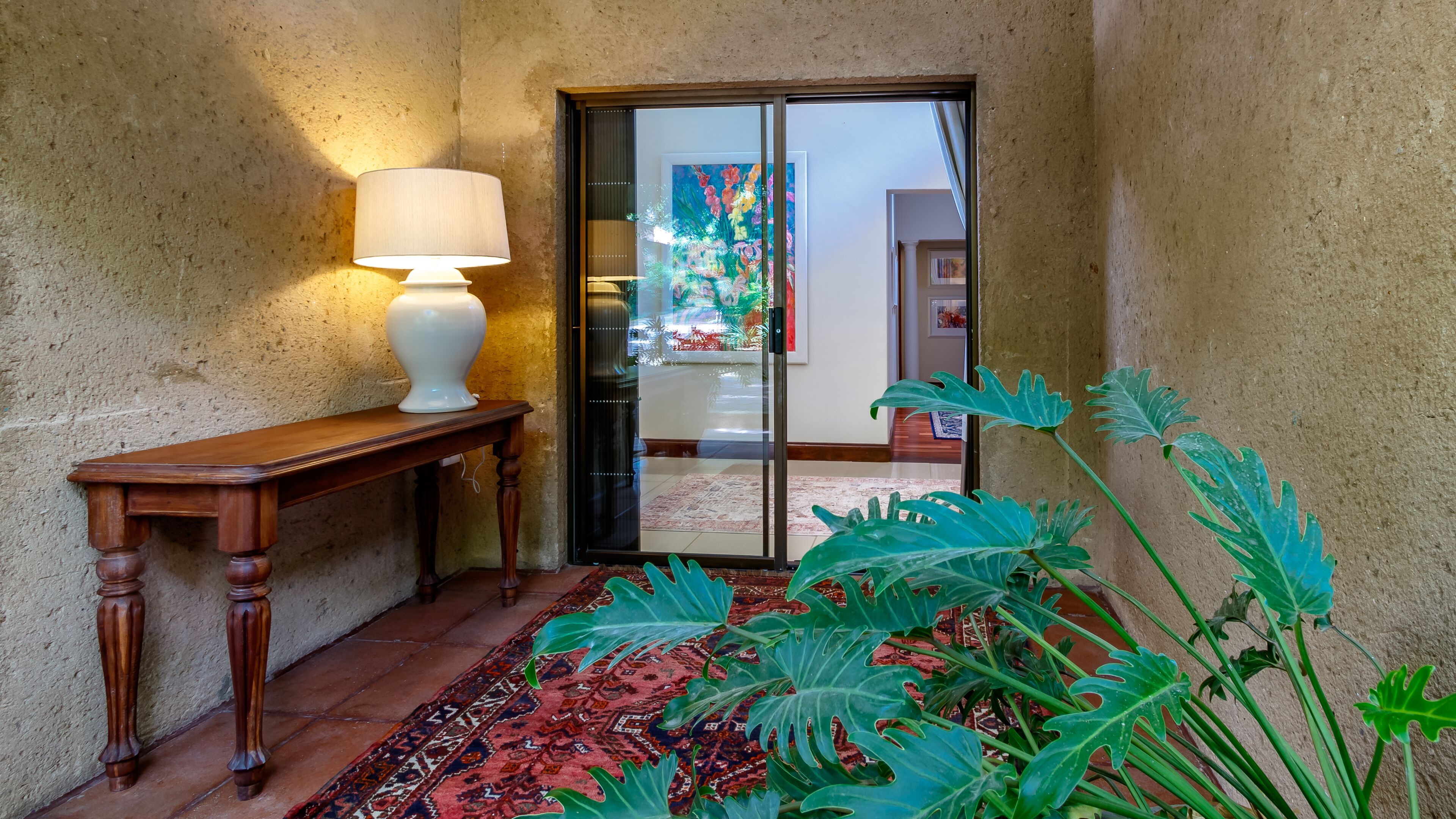Un pasillo con una mesa de madera y una lámpara, un vidrio que da a una habitación con un cuadro colorido, y plantas verdes grandes.