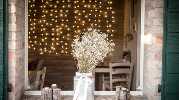 Une entrée rustique avec une porte en bois, un vase en métal contenant des fleurs et un mur de briques décoré de guirlandes lumineuses.