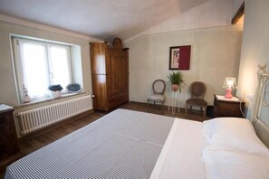 Un dormitorio con una cama grande, un armario de madera, un radiador, una ventana con cortinas, una silla, una mesita de noche con una lámpara y un cuadro en la pared.