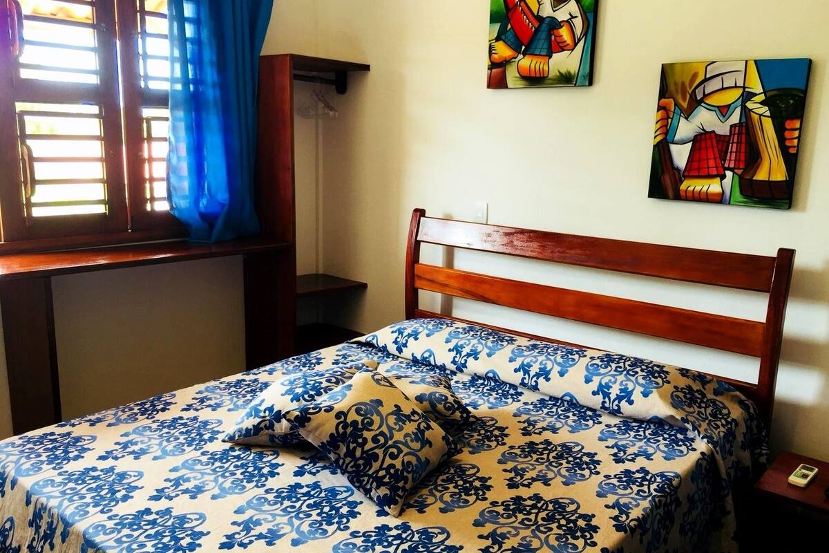 A bedroom with a wooden bed frame, a patterned bedspread, and two pillows. There is a window with blue curtains, a bedside table, and two artworks on the wall.