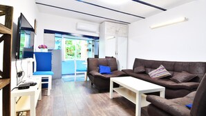 Uma sala de estar moderna com um sofá de couro marrom, uma mesa de centro branca e uma poltrona azul. Há uma televisão sobre um suporte de madeira, e um armário com porta branca.
