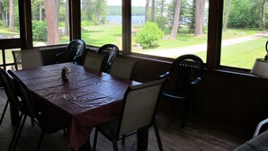 Un área de comedor cubierta con una mesa dispuesta para seis personas, rodeada de sillas y grandes ventanales con vistas a un bosque y un lago.