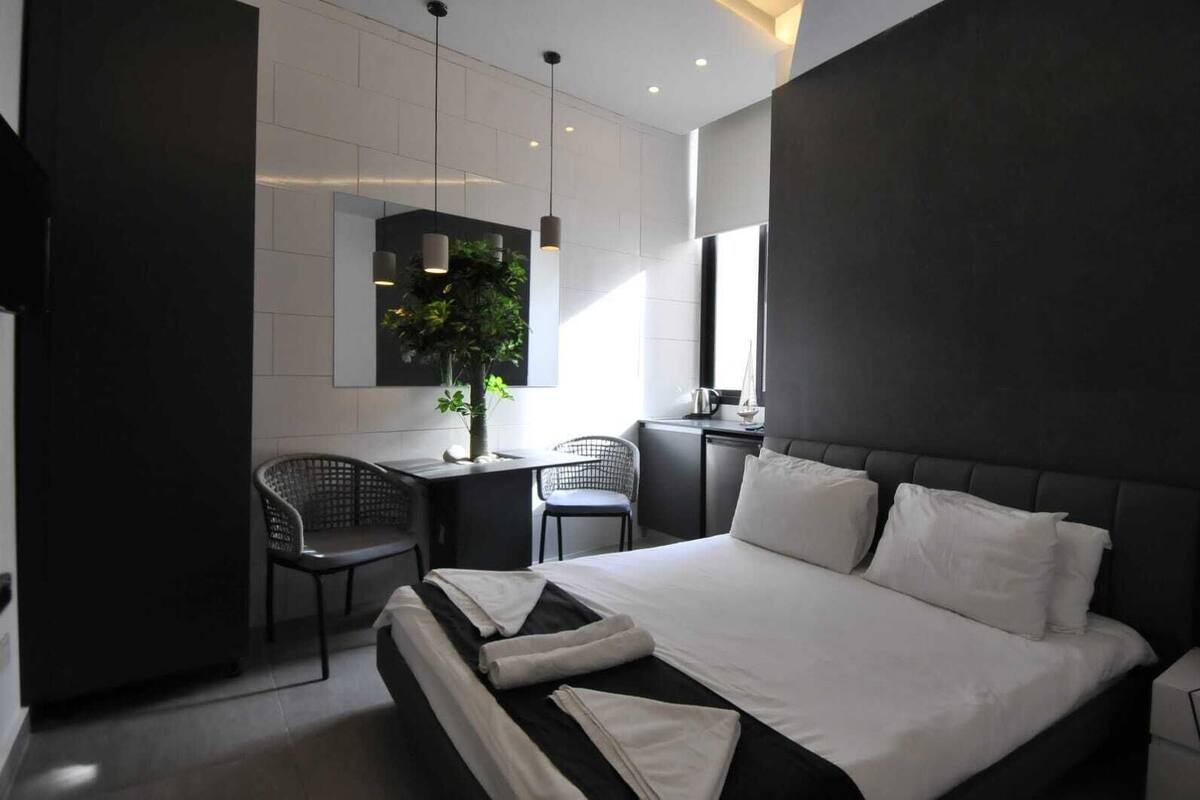 Une chambre d’hôtel moderne avec un grand lit, un bureau avec deux chaises, une plante en pot et un grand miroir au mur.