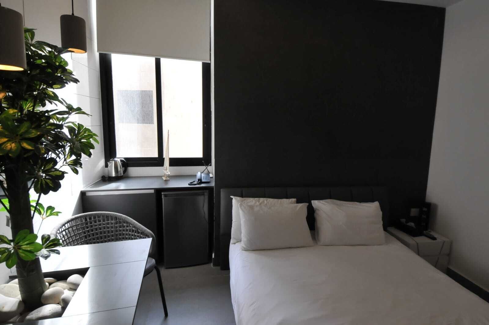 A modern hotel room with a bed, a desk, and a chair. There is a window providing natural light, and a plant is visible.