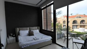 A modern bedroom with a large bed, a small bedside table, and a view of a courtyard with outdoor seating. - Batroun Bahsa Bay (Batroun)