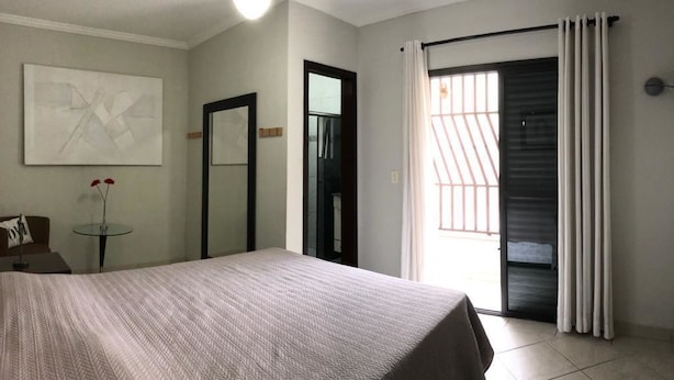 Habitación de hotel con cama, ventilador de techo, puerta, ventana con cortinas y una pequeña mesa con un florero.