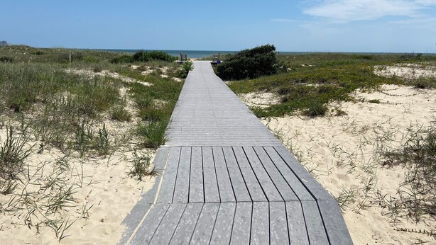 Una pasarela de madera que conduce a la playa, rodeada de dunas de arena y vegetación escasa.