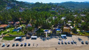 An aerial view of a beach resort with rows of sun loungers and umbrellas, a swimming pool, and a backdrop of palm trees and buildings. - Entire Palmar · Stunning 4 BR Oceanfront Townhome, amazing views. (Sayulita)