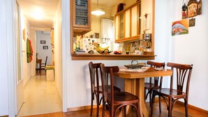 A dining area with a wooden table and four chairs, a kitchen with wooden cabinets and various appliances, and a hallway leading to another room. - Happy place at the foot of Acropolis. (Athens)