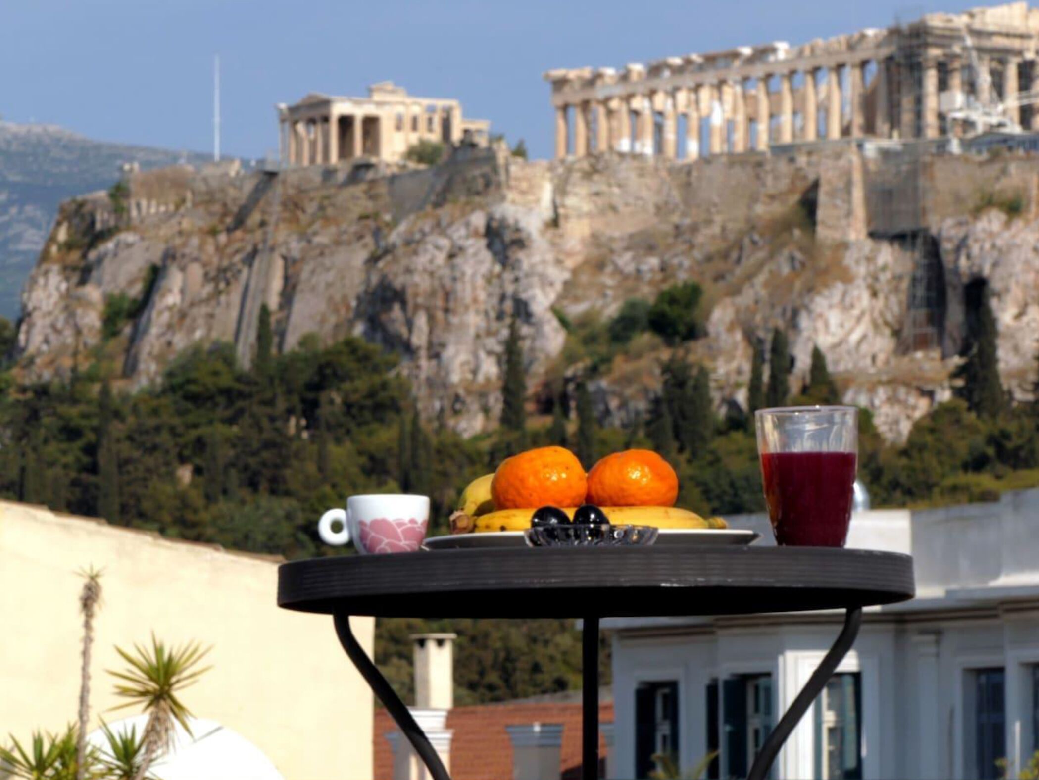 A table with fruits, a cup, and a glass of red drink, overlooking ancient ruins.