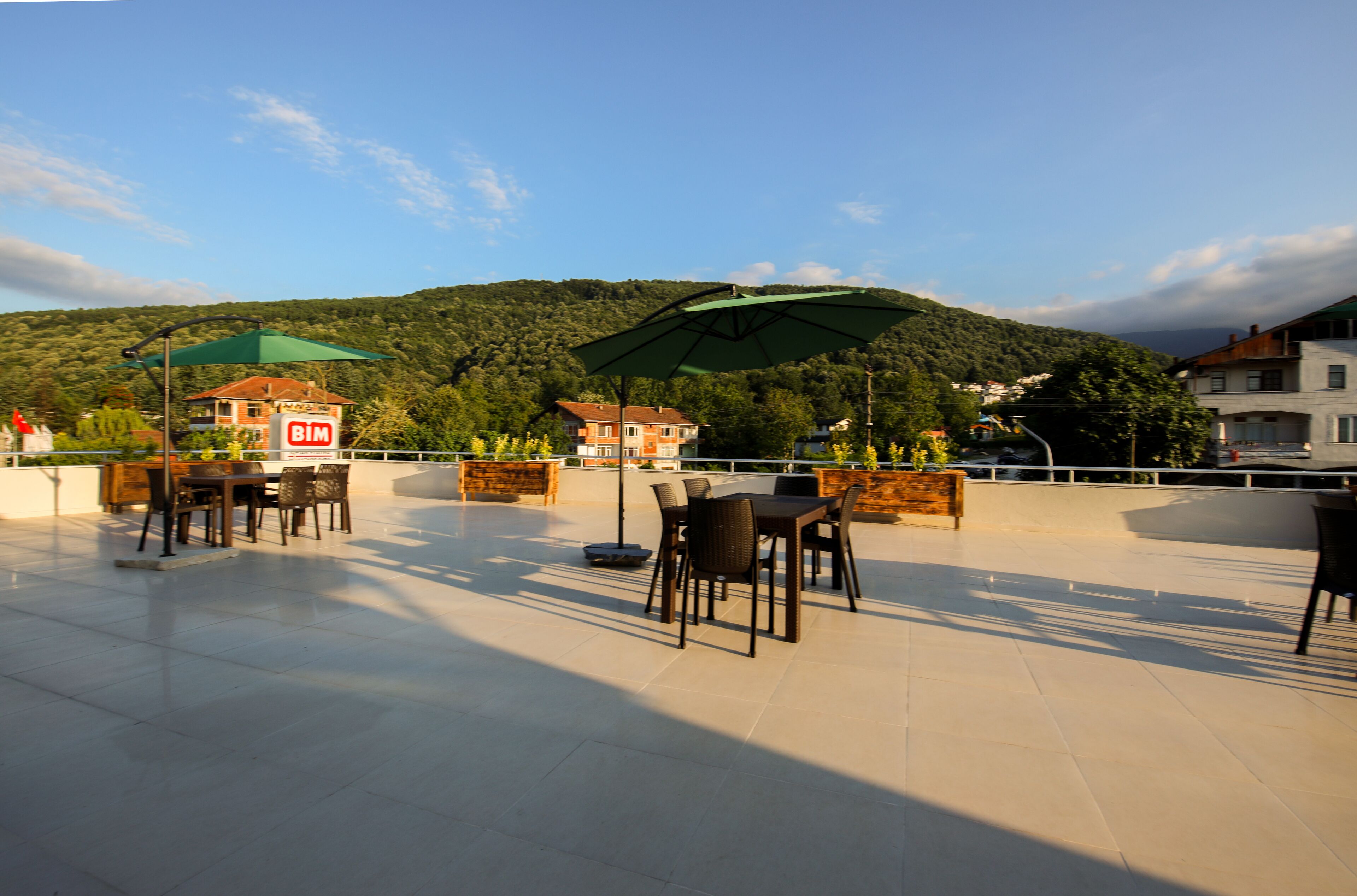 Una terrazza esterna con tavoli e sedie, un ombrellone verde e vista su una collina con edifici sullo sfondo.