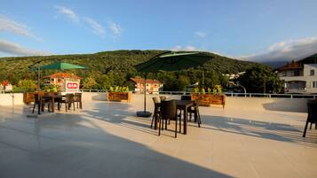 Una terrazza esterna con tavoli e sedie, un ombrellone verde e vista su una collina con edifici sullo sfondo.