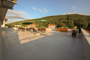 An outdoor terrace with tables and chairs, umbrellas, and a view of a forested hill.